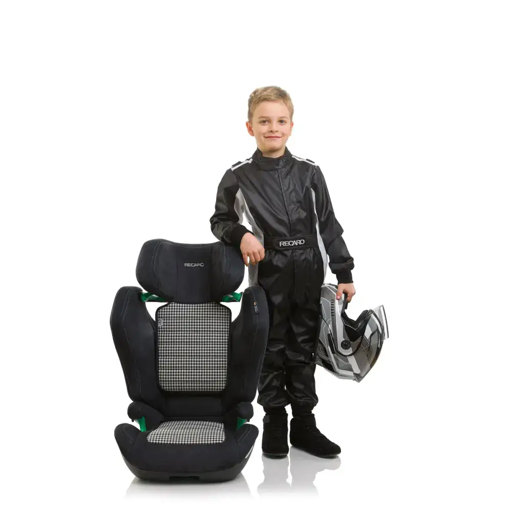 Child in a black racing suit holding a helmet beside the Recaro AXION 1 Pepita booster seat.