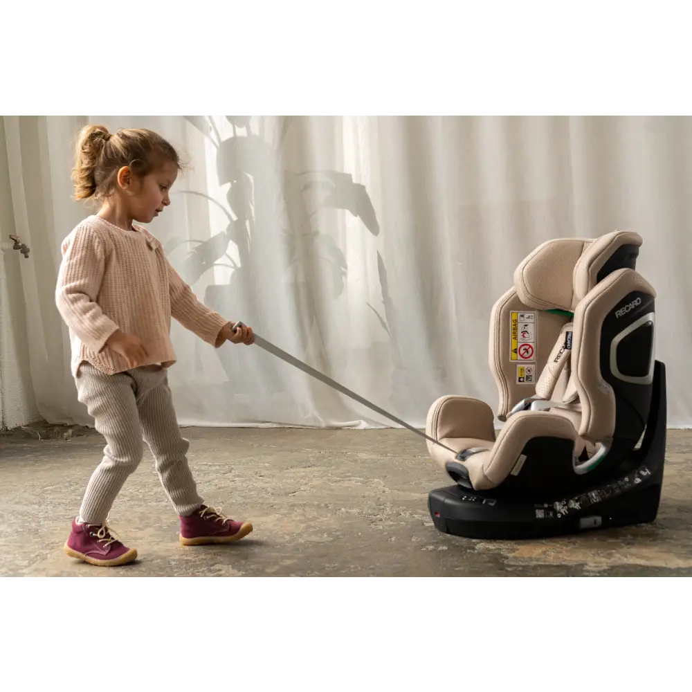 Child holding the harness adjustment strap of an Elegant Beige XENON seat on a concrete floor.