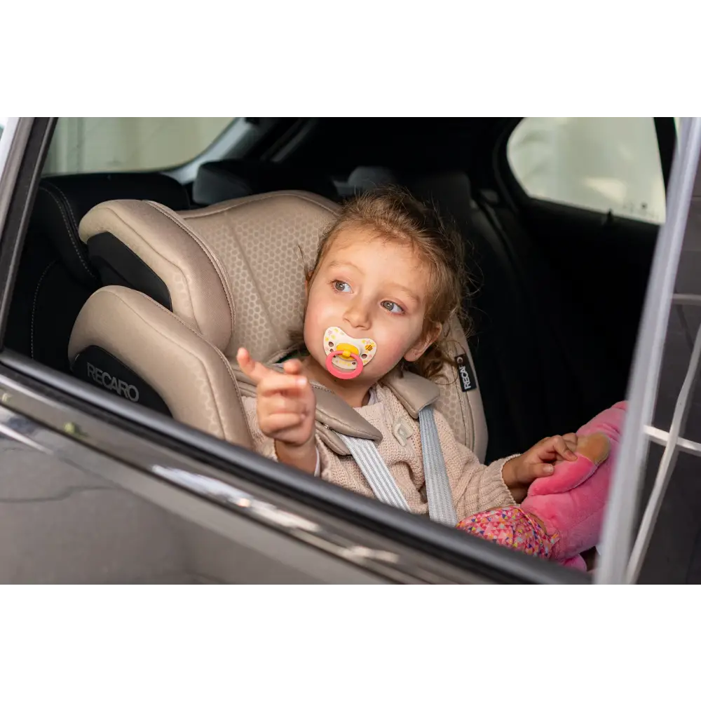 Child with a dummy wearing the padded harness in an Elegant Beige XENON, viewed through an open vehicle window.