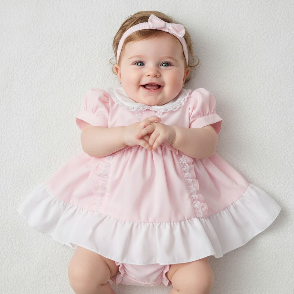 A baby girl wearing a soft pink and white ruffled dress with lace trim, headband, and pants set