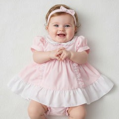 A baby girl wearing a soft pink and white ruffled dress with lace trim, headband, and pants set