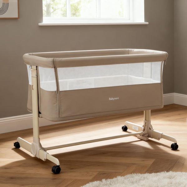 Beige babymore neera bedside crib with mesh sides and black caster wheels for easy mobility and ventilation