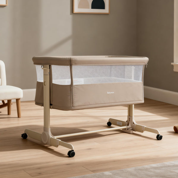 Beige babymore neera bedside convertible crib with mesh sides and caster wheels for easy mobility