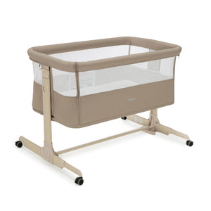 Beige babymore neera bedside crib convertible baby bassinet with clear mesh sides and black caster wheels