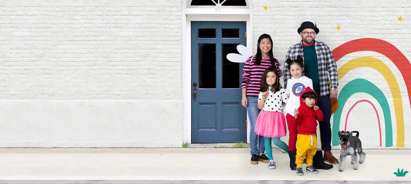 A family of five and a small dog stand together in front of a white brick house with a blue door, positioned beside a colorful rainbow mural painted on the wall.