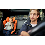Child holding a soft toy in a Vibrant Orange AXION 1 on the rear seat, with an adult at the wheel.
