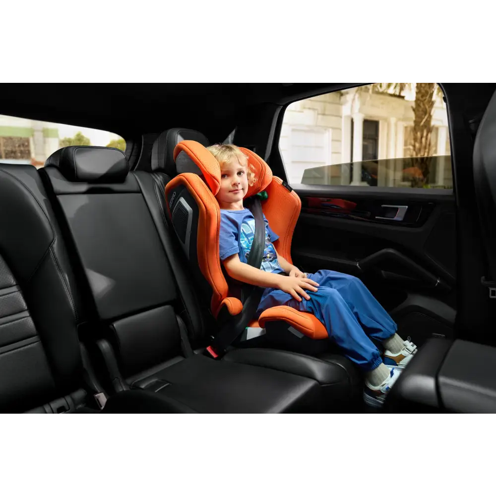 Child seated in a Vibrant Orange AXION 1 in a vehicle, wearing a diagonal seat belt.